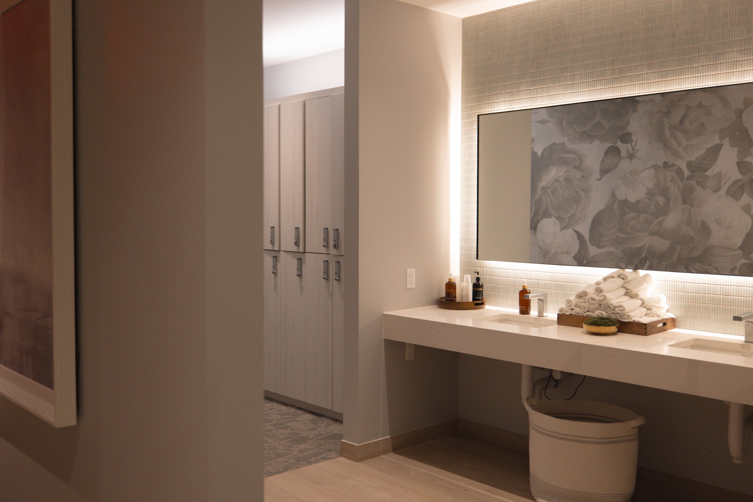 Spa locker room with modern cabinetry, a clean vanity featuring towels and skincare products, illuminated mirror, and floral artwork, reflecting Exhale Spa's tranquil wellness environment.