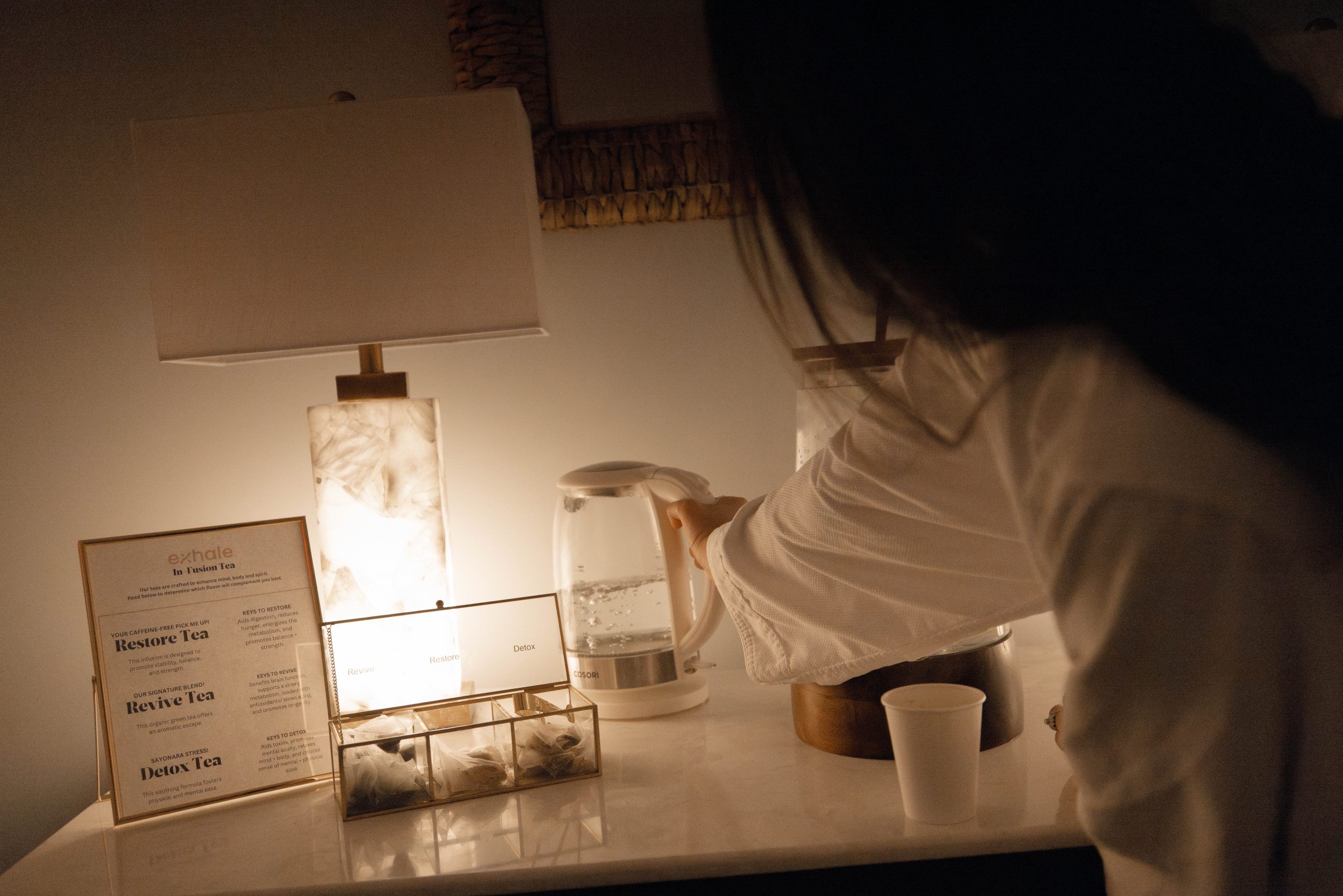 Person in a spa robe preparing herbal tea in a serene wellness environment, featuring a glowing lamp, a tea selection box with "Restore," "Revive," and "Detox" labels, and a glass kettle, emphasizing relaxation and rejuvenation.