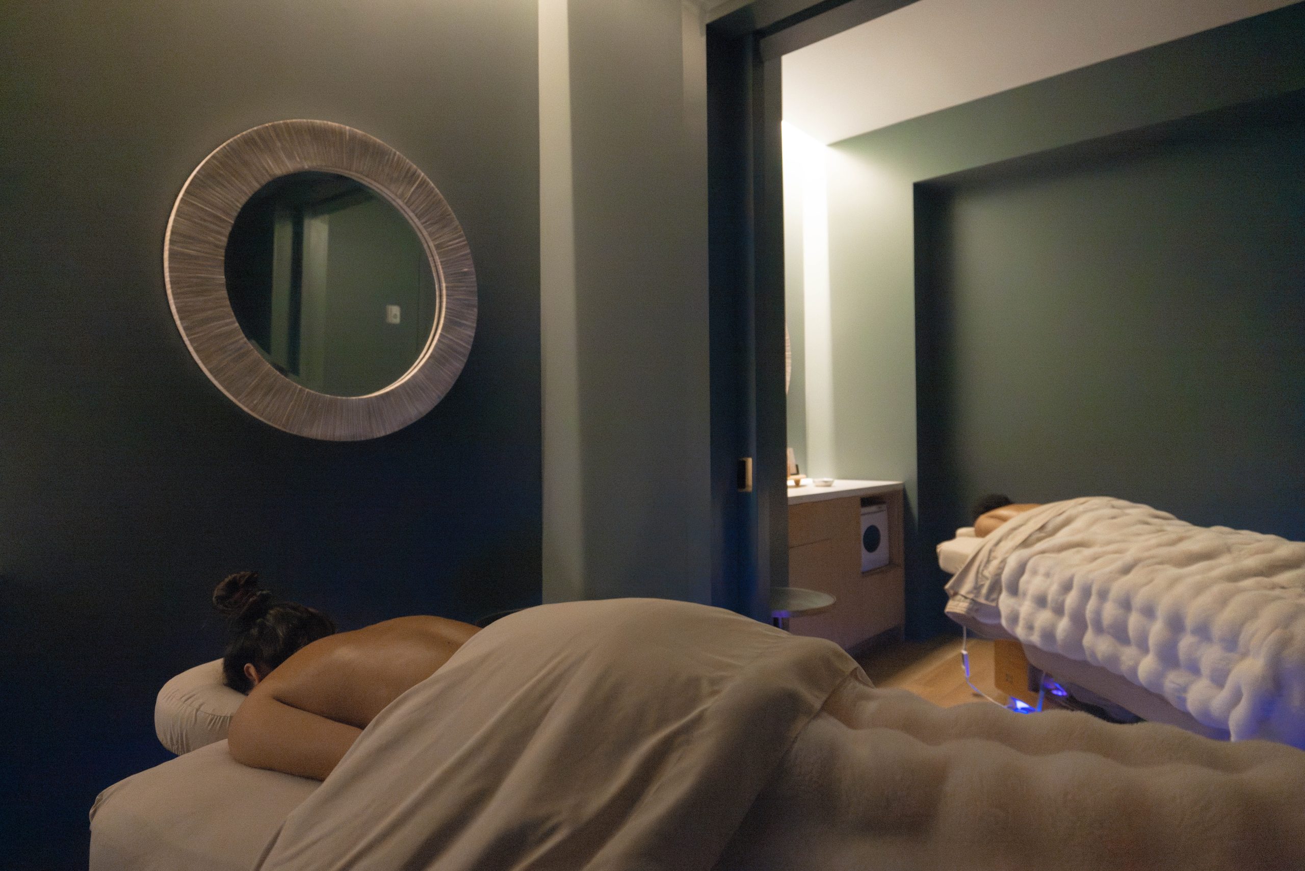 Spa treatment room with two massage tables, one occupied by a person, and a decorative mirror on the wall, highlighting a serene wellness environment at Exhale Spa in Central Park.