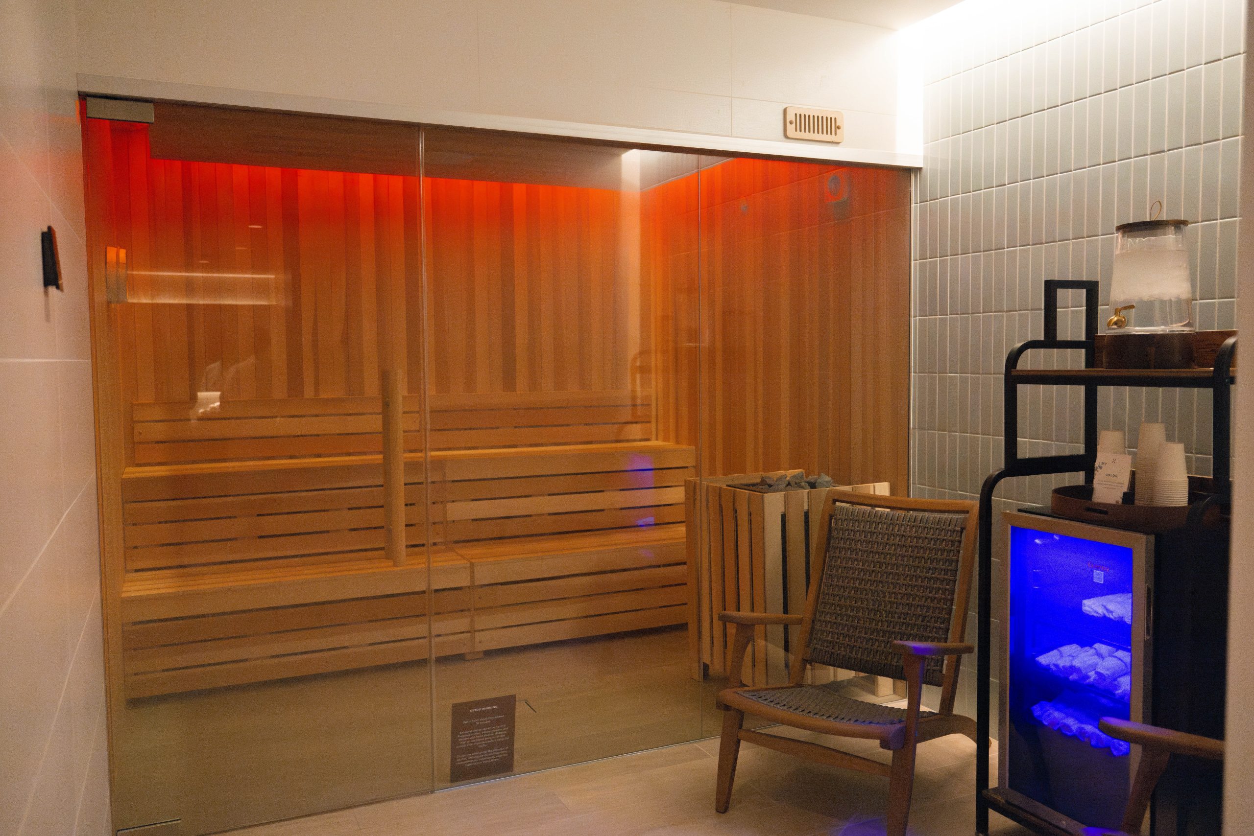Spa sauna room with wooden benches, glass door, ambient red lighting, and a chair beside a hydration station.