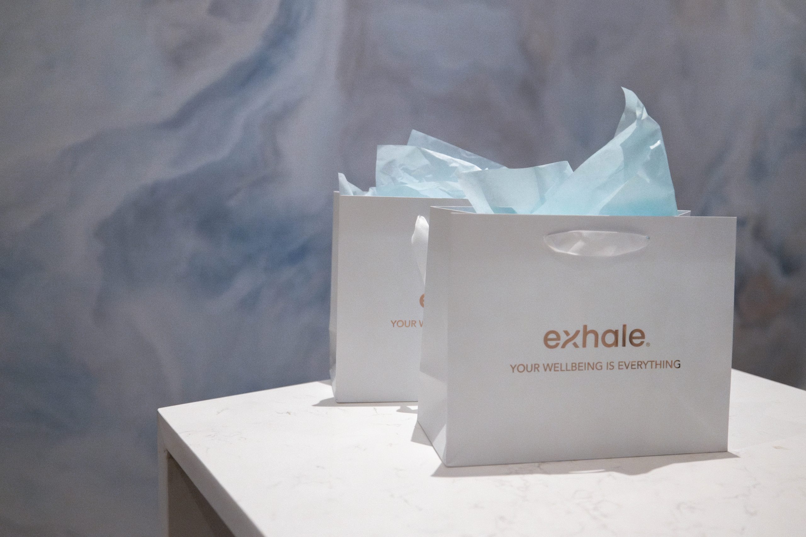 Exhale Spa gift bags on a marble table with blue tissue paper, featuring the tagline "YOUR WELLBEING IS EVERYTHING" in a serene wellness environment.