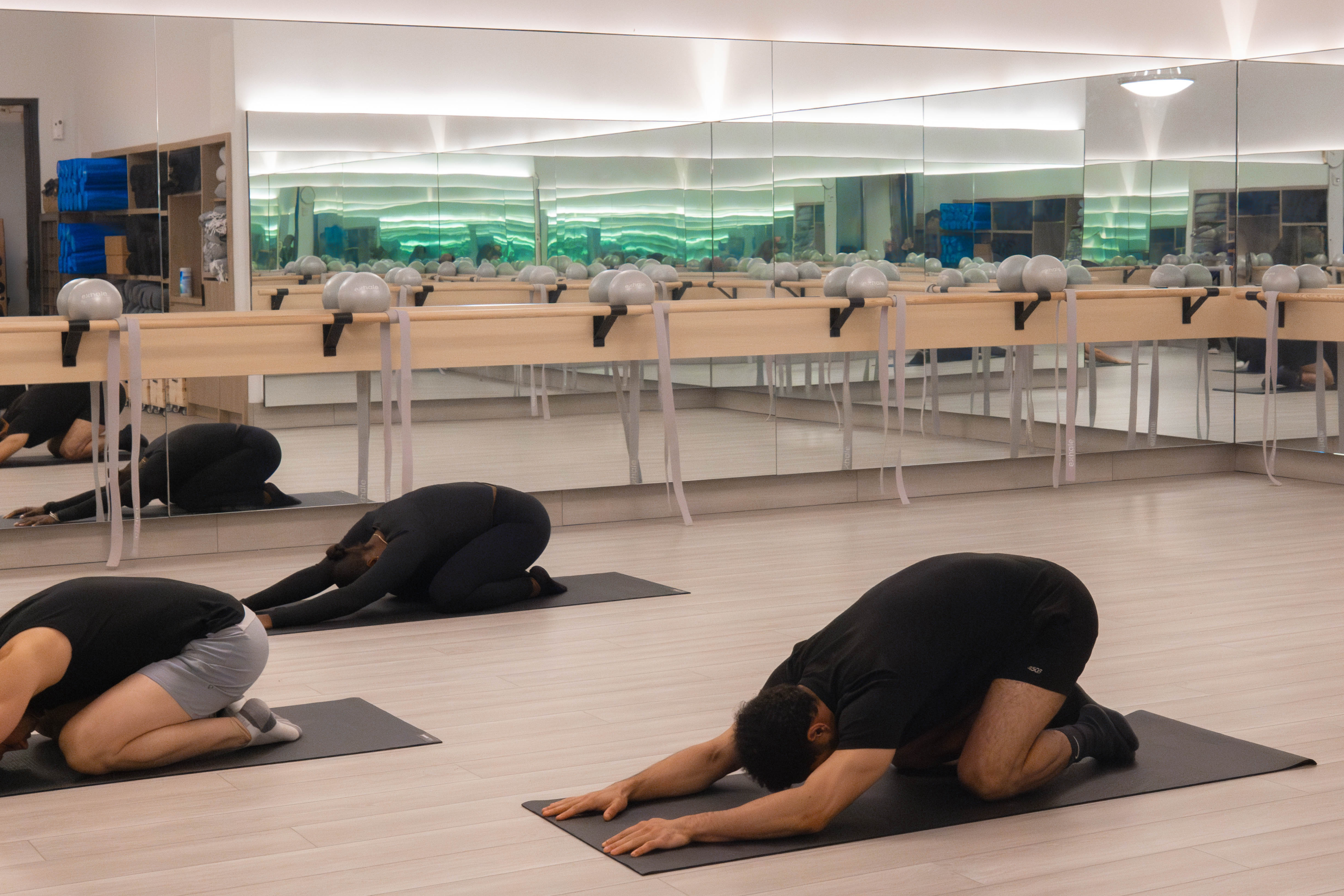 Individuals practicing yoga stretches in a well-lit fitness studio with mirrors and exercise equipment, emphasizing wellness and fitness classes at Exhale Spa.
