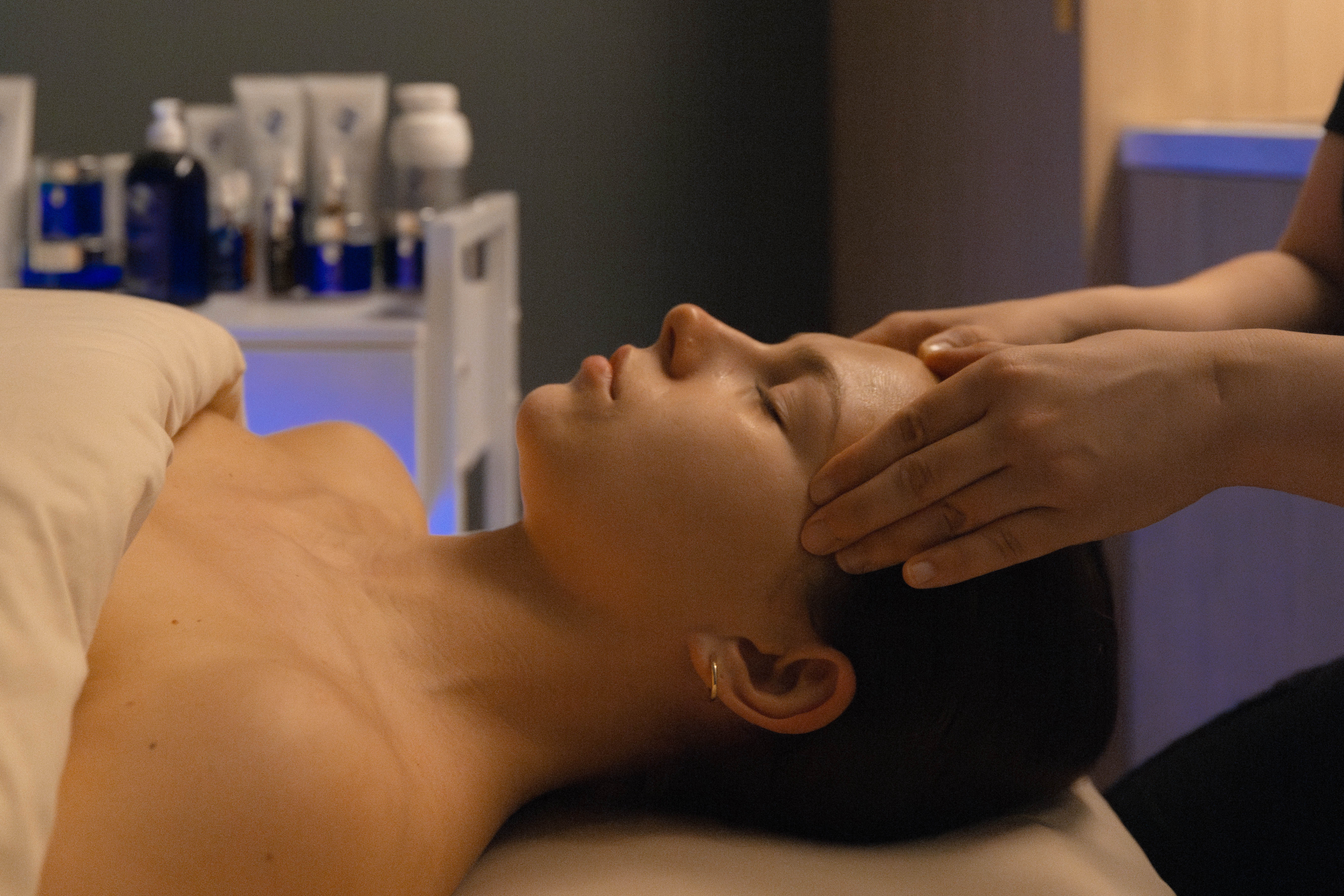 Woman receiving a facial treatment in a spa setting, with skincare products visible in the background, highlighting wellness and relaxation services at Exhale Spa.