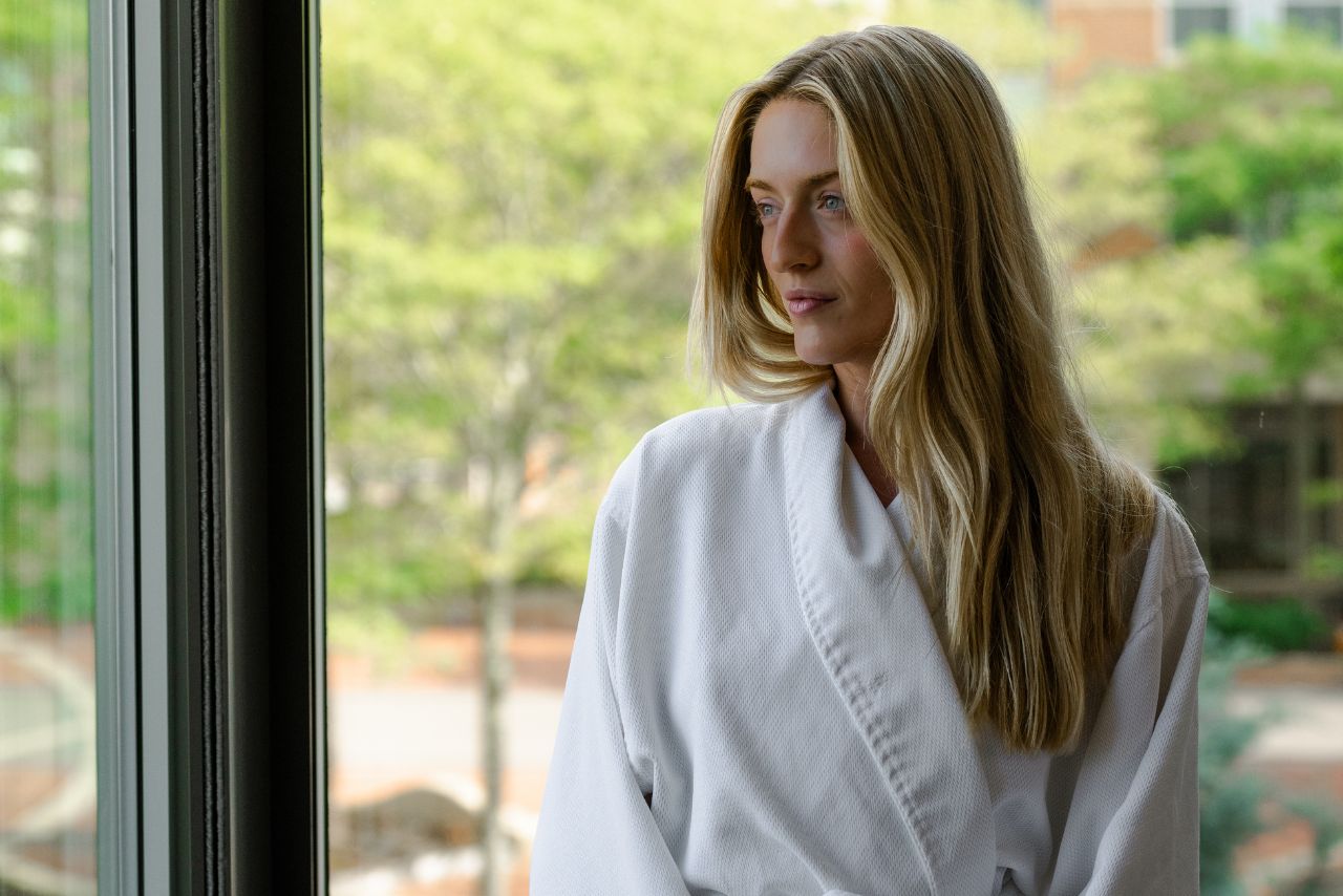 Woman in a white spa robe gazing out of a window, surrounded by greenery, representing relaxation and wellness at Exhale Spa in Battery Wharf, Boston.