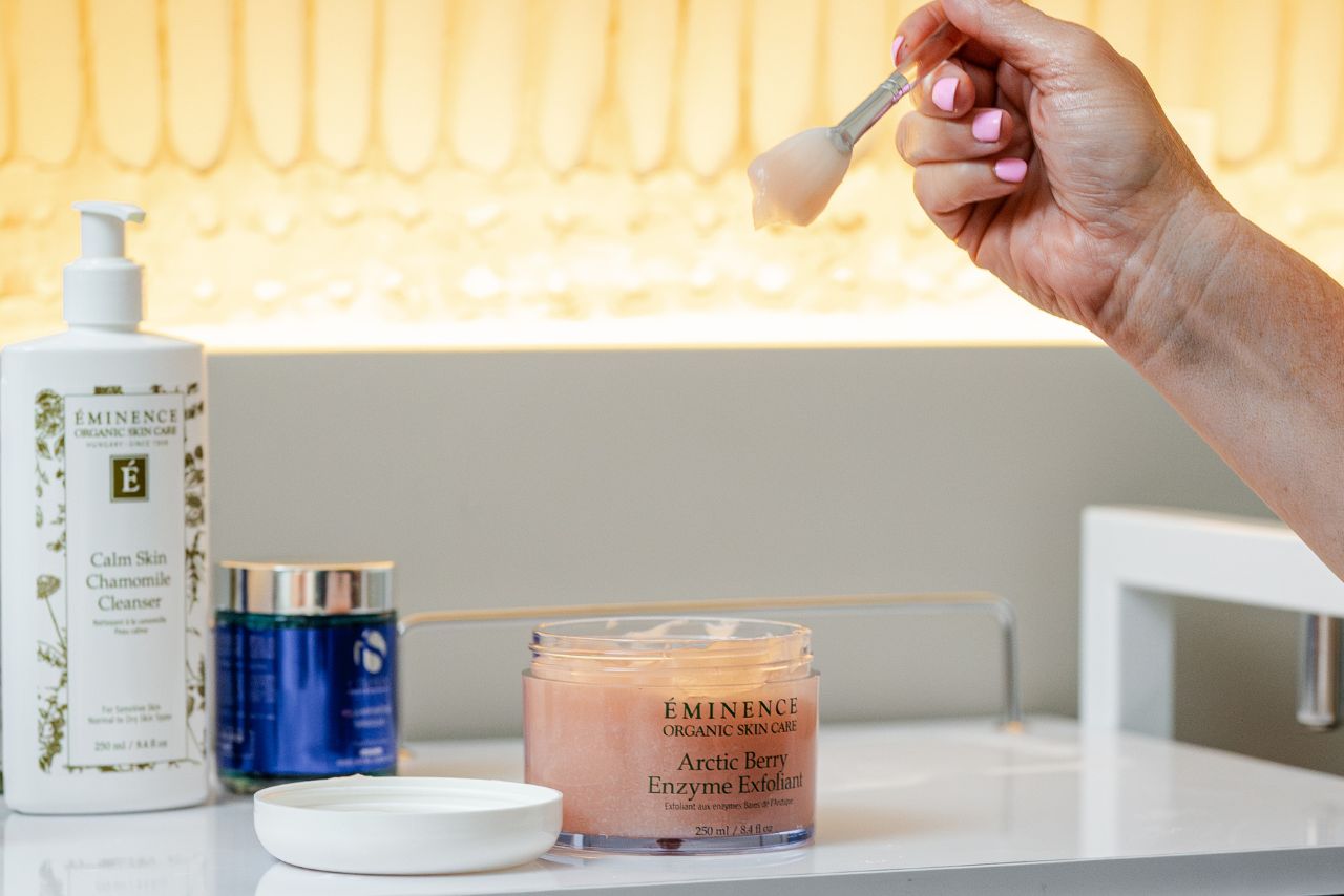 Hand holding a brush with Arctic Berry Enzyme Exfoliant from Éminence Organic Skin Care, Calm Skin Chamomile Cleanser and a blue skincare product in the background, showcasing spa treatments at Exhale Spa in Battery Wharf, Boston.